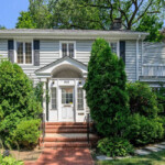 two story house with trees in the background and bushes in front